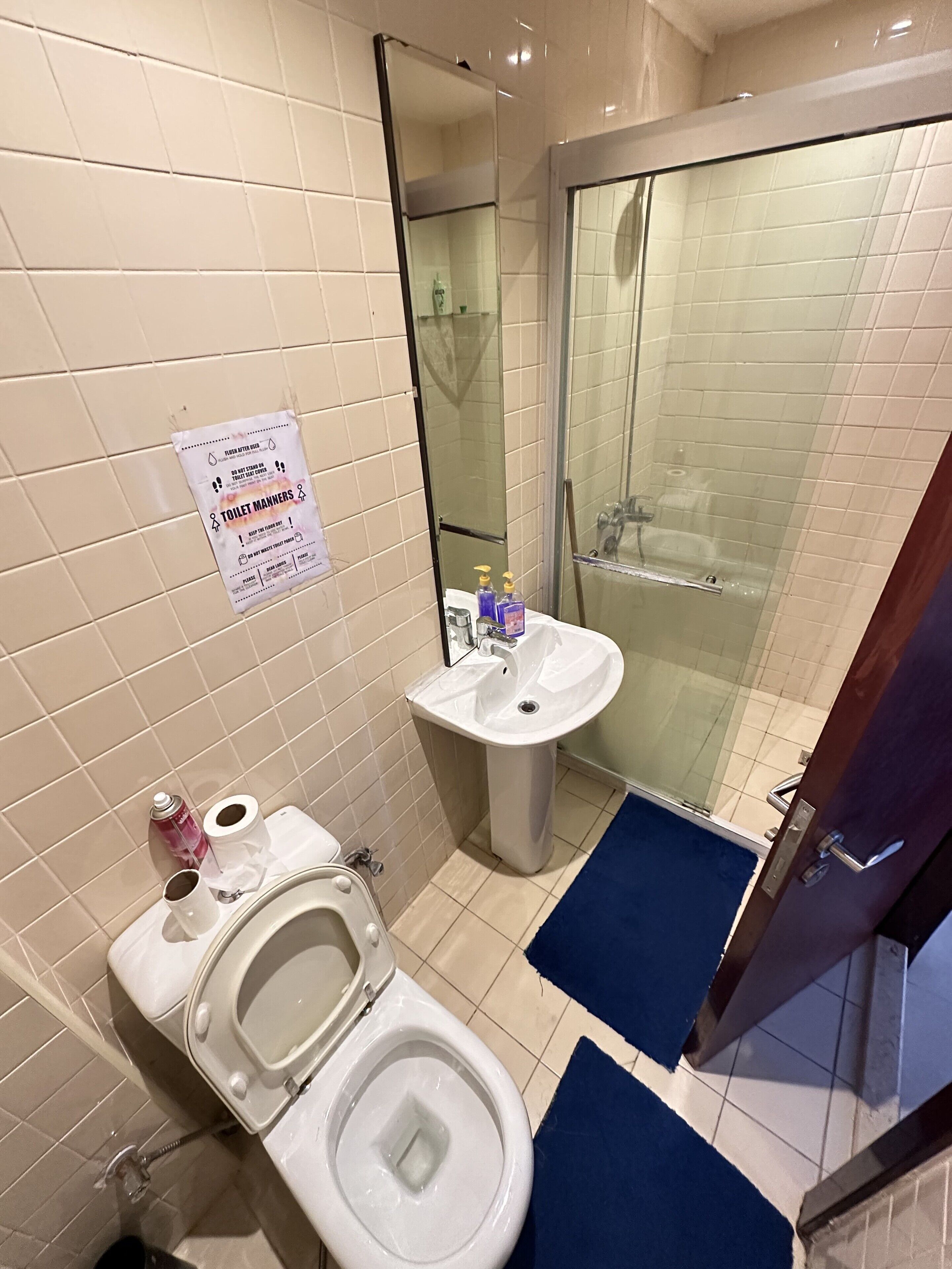 A bathroom with a toilet, sink, and shower. There is a sign on the wall and a blue mat on the floor.