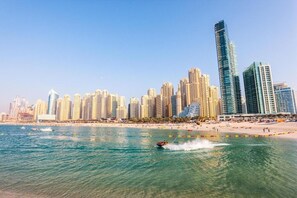 A coastal cityscape with high-rise buildings, a beach, and a jet ski in the water. - Bollywood Beach Hostel (Dubai)