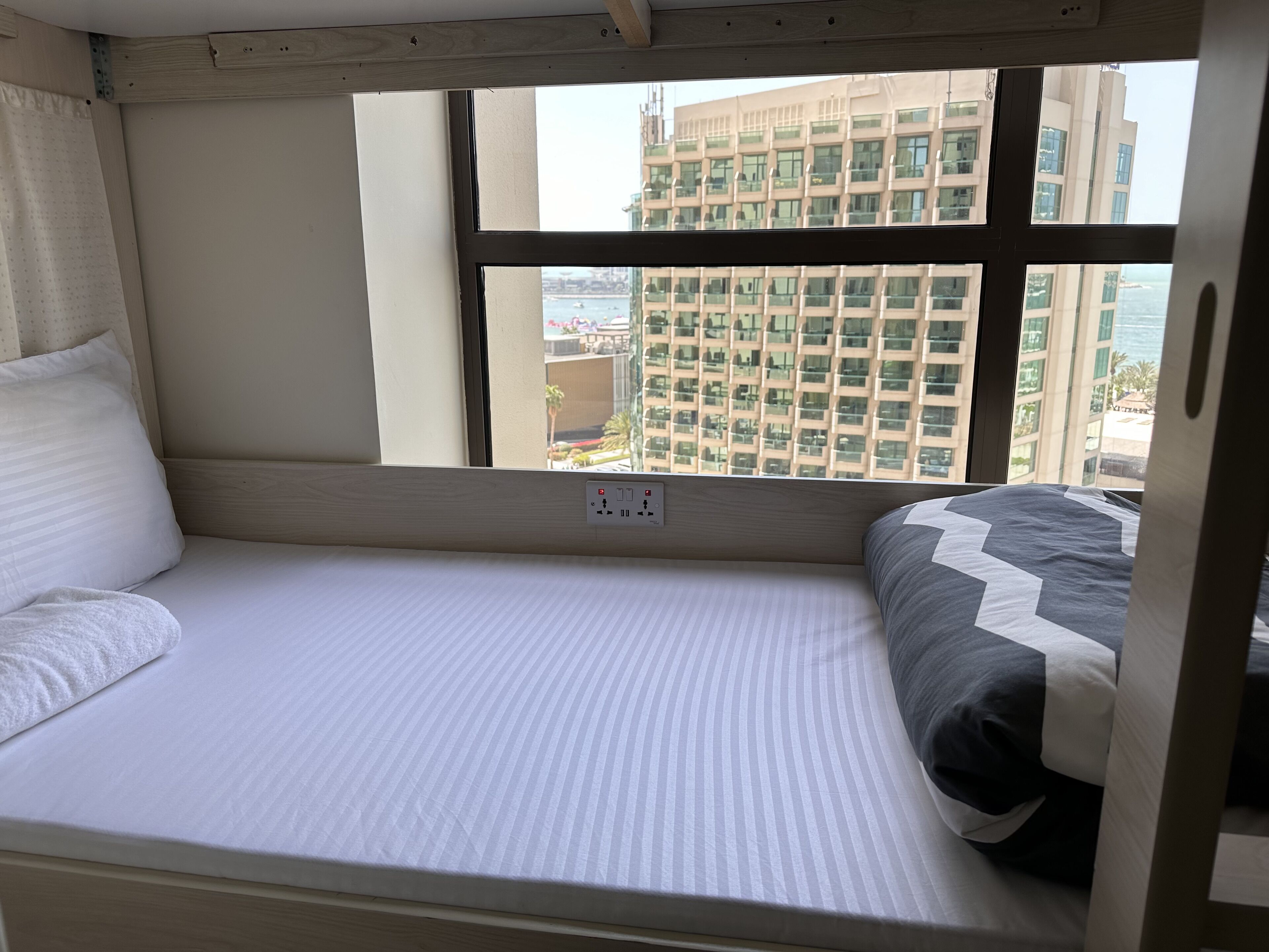 A single bed with a striped mattress, a pillow, and a blanket. There is a window with a view of a building and a body of water.