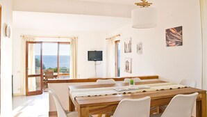 Een eetruimte met een houten tafel, witte stoelen en uitzicht op zee door grote ramen.