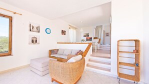 Een ruime zithoek met een rieten bank, een witte zacht beklede bank en een rieten bijzettafeltje. Een trap leidt naar een eetgedeelte met een witte tafel en stoelen.