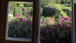 Una ventana con vistas a un paisaje montañoso y un jardín cuidado con flores púrpuras.