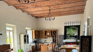 Amplia sala de estar con un sofá, un banco a rayas y una mesa de comedor con sillas. Se observa una zona de cocina con armarios de madera y un ventanal grande.