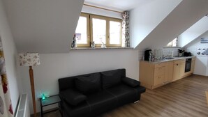 A modern apartment with a black leather sofa, wooden flooring, a kitchenette, and a large window with floral curtains. - Cozy vacation apartment, (Füssen)