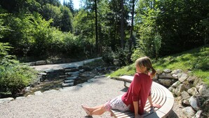 A woman sitting on a bench in a park with a stream and trees in the background. - Cozy vacation apartment, (Füssen)
