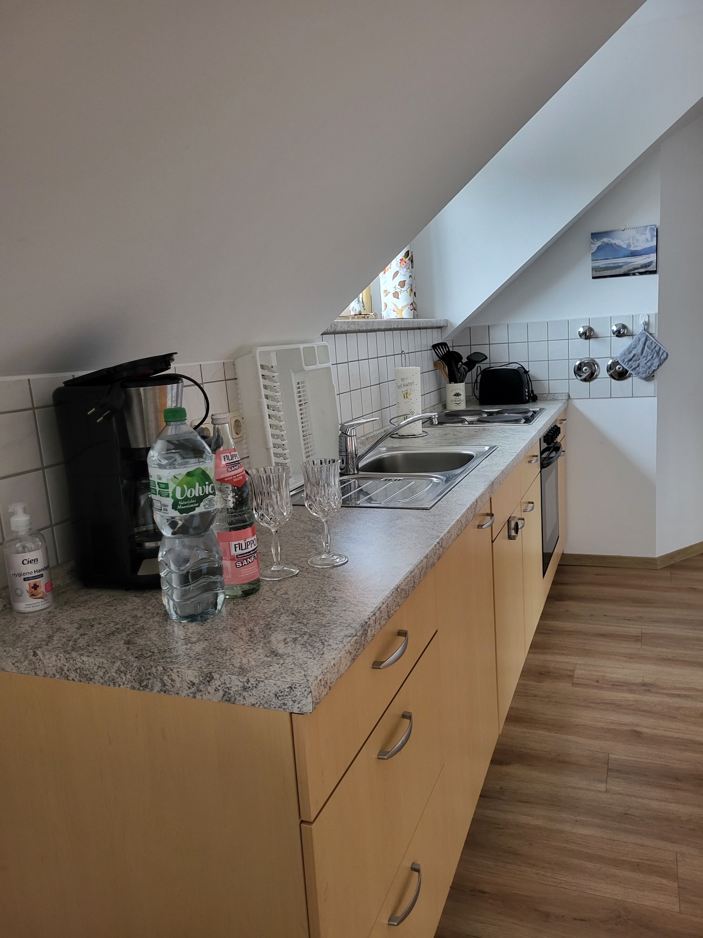 A kitchen with a granite countertop, a coffee maker, a water dispenser, a bottle of hand sanitizer, and a wine glass.