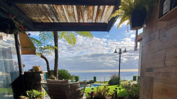 Eine Holzterrasse mit einem Korbstuhl, einem Pflanzgefäß aus Stein mit Blumen und Blick aufs Meer.