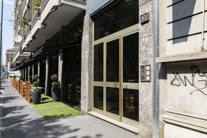 Un edificio con grandes puertas de cristal, fachada de piedra y una pequeña zona ajardinada con plantas en macetas.