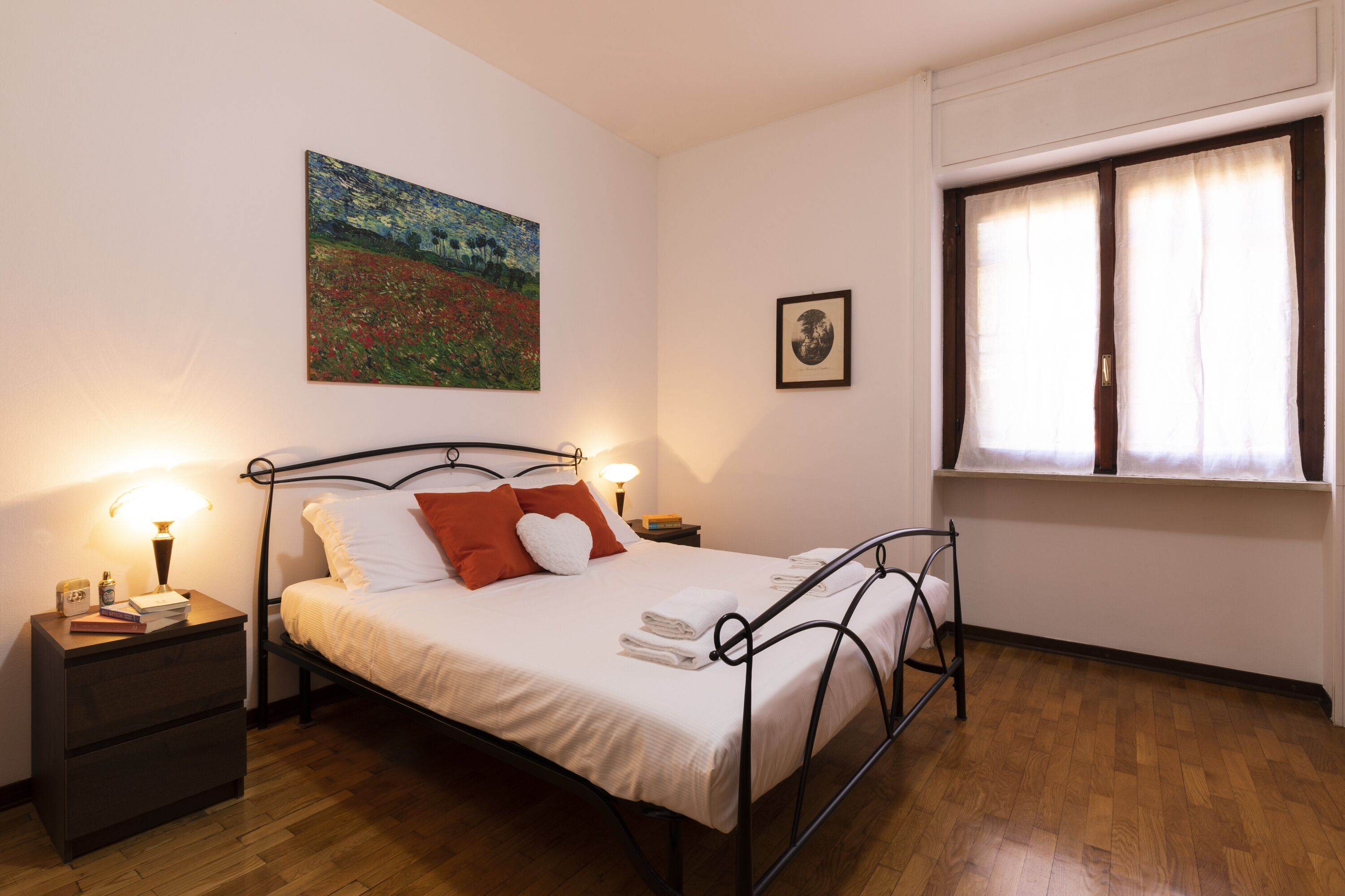 A bedroom with a metal bed frame, white bedding, a painting on the wall, a bedside table with a lamp, and a window with curtains.