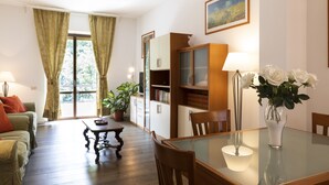 A living room with a sofa, coffee table, and dining table with chairs. There is a bookshelf, a potted plant, and a vase with flowers.