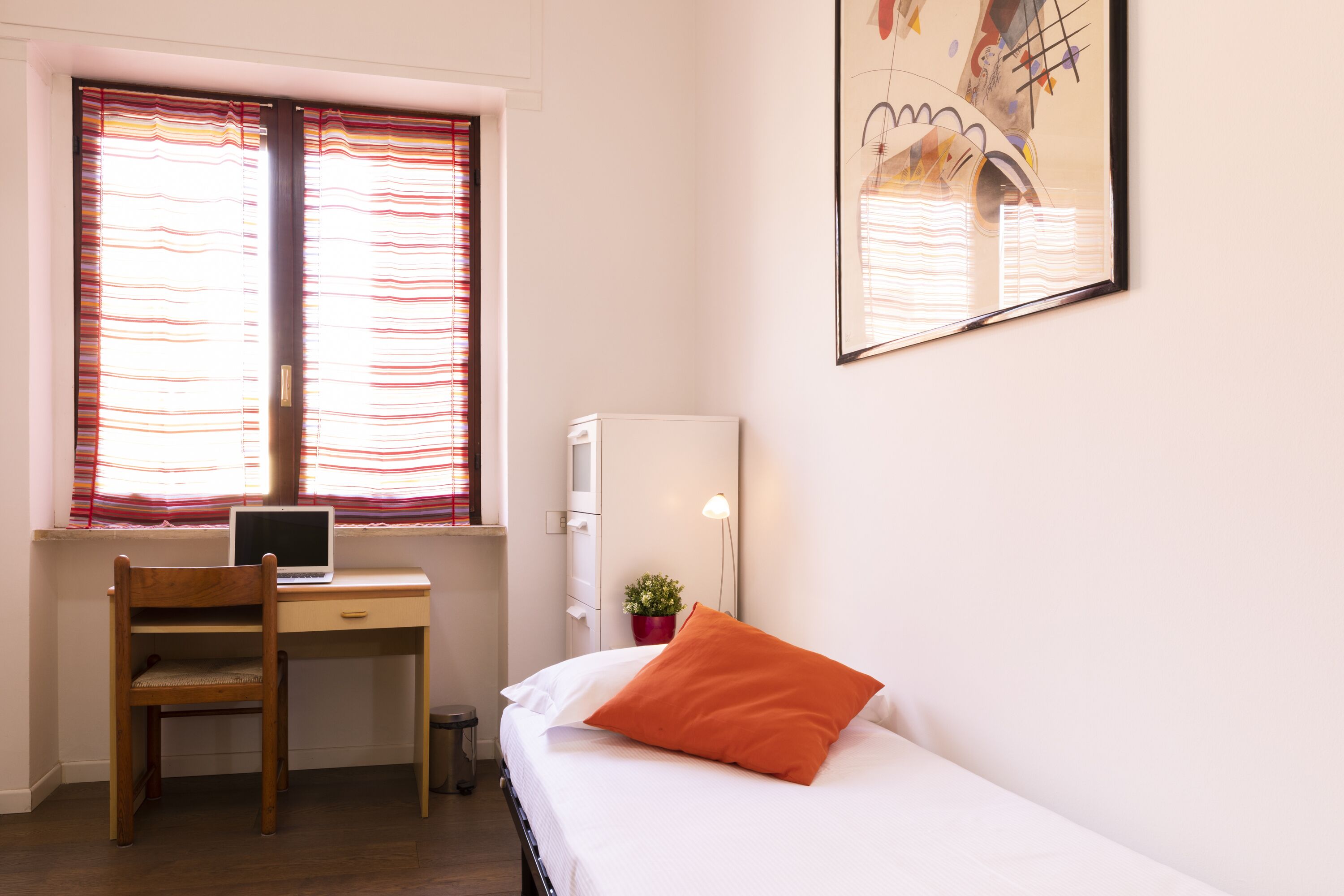 A bedroom with a bed, a desk, a chair, a window with striped curtains, a lamp, and a small plant.