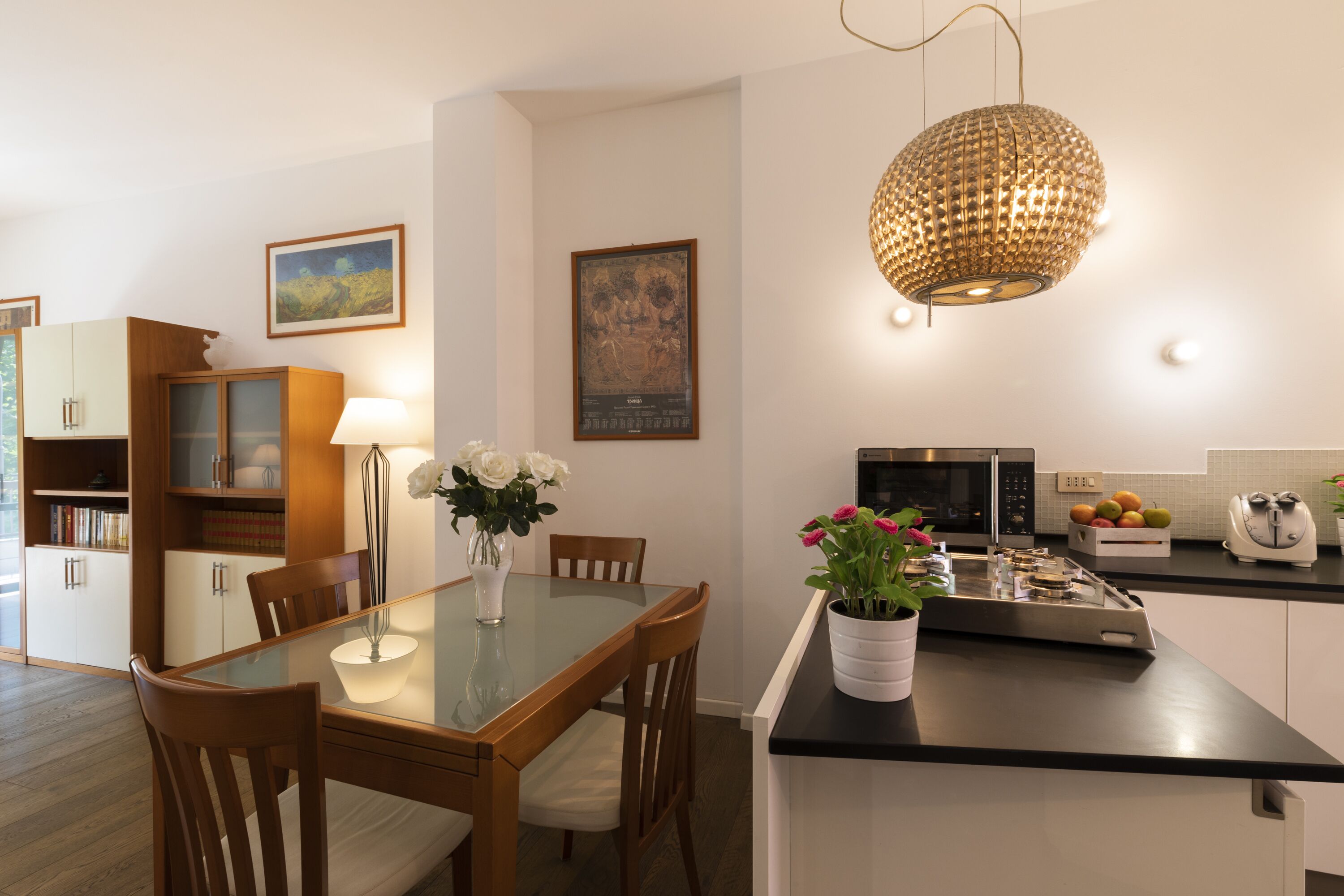 A modern kitchen with a dining area, featuring a glass-top table, wooden chairs, a white cabinet, a microwave, and a fruit basket.