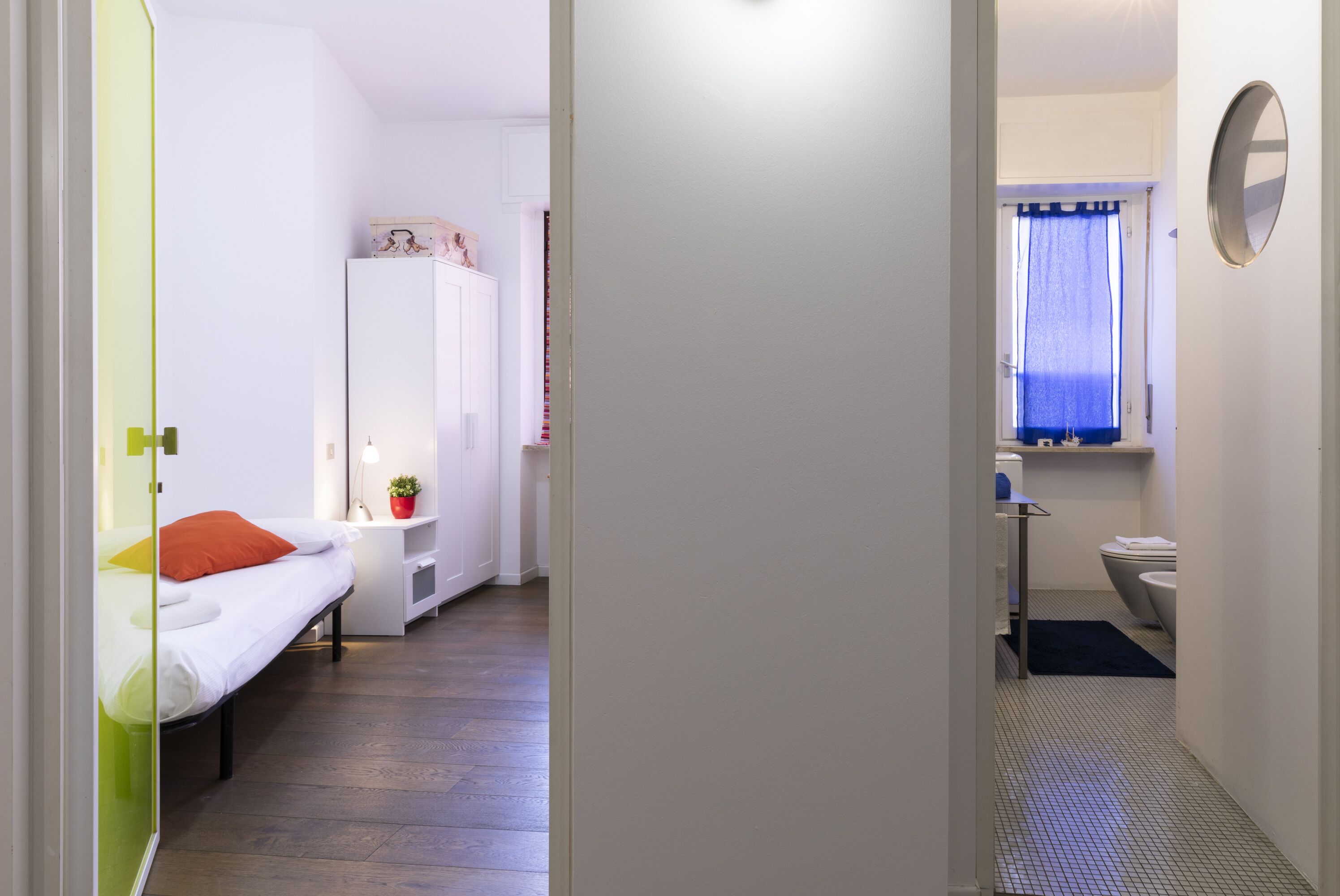Ein kleines Zimmer mit Bett, Kleiderschrank, einem kleinen Tisch mit Pflanze sowie einem Badezimmer mit Toilette und Waschbecken.