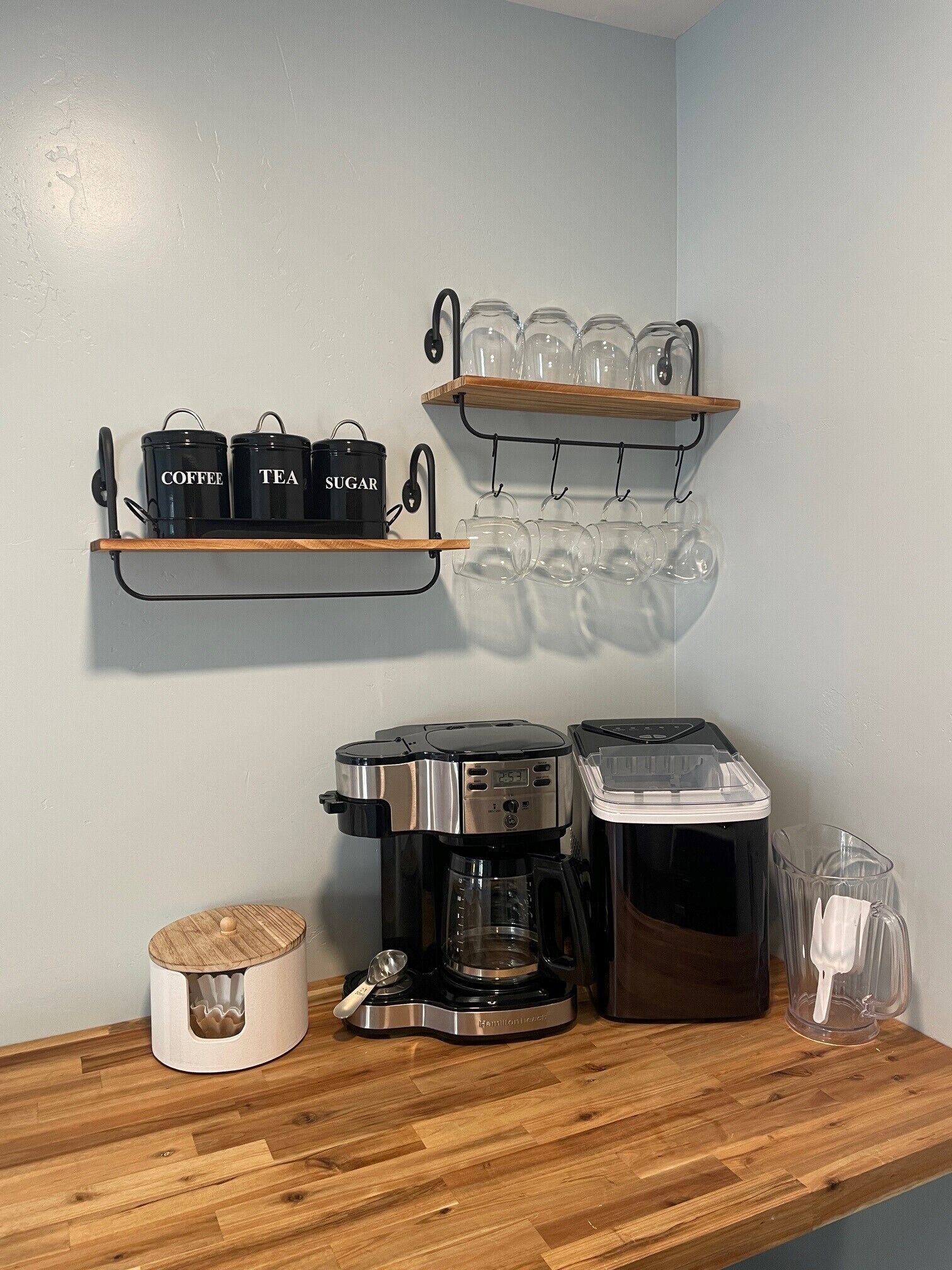 A kitchen area with a coffee maker, a rice cooker, and a blender on the counter. There are shelves with jars labeled 'coffee', 'tea', and 'sugar', and a rack with glasses.