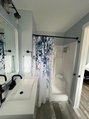 A bathroom with a white sink, a mirror, a shower with a floral curtain, and a door leading to another room.