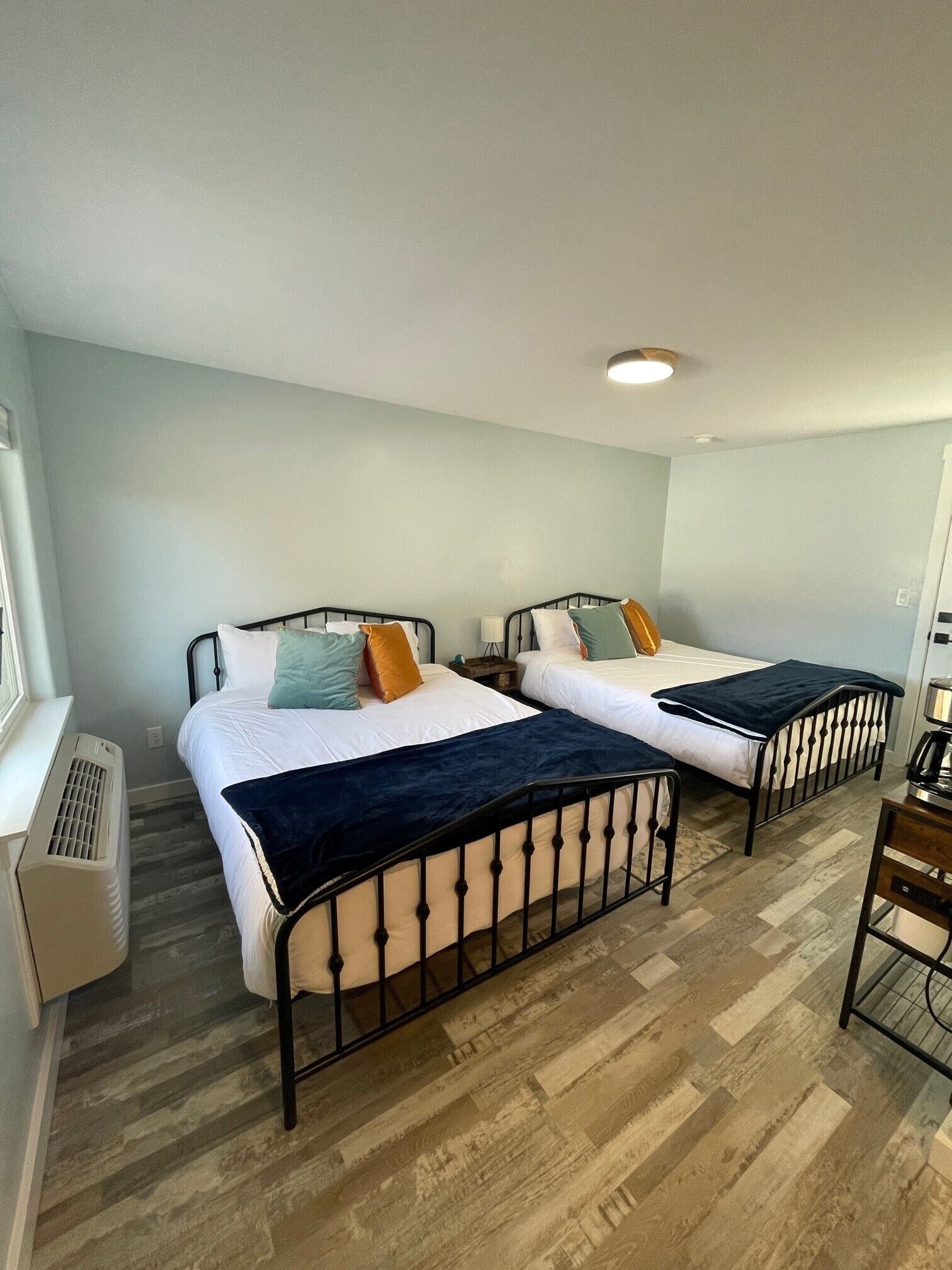 A room with two beds, each with a black metal frame and white bedding. There is a heater on the left and a desk with a chair on the right.