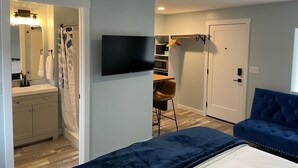 Une chambre moderne avec un grand lit, un téléviseur fixé au mur, une salle de bain avec un rideau de douche et un petit coin salon avec un fauteuil.