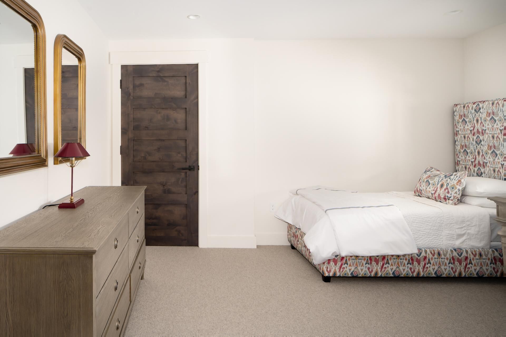 Une chambre avec une commode en bois, un lit avec une literie à motifs, un miroir et une porte.
