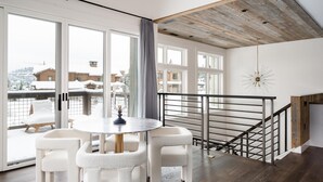 Una sala de estar moderna con una mesa redonda y cuatro sillas blancas, una escalera de madera y ventanas amplias que dan a un paisaje nevado.