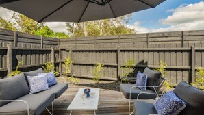 Un patio exterior con terraza de madera, dos sofás grises con cojines estampados, una mesa blanca con dos vasos y un paraguas grande que da sombra.