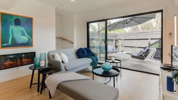 Una sala de estar moderna con un sofá gris, una chimenea y un amplio ventanal corredera que da a un patio exterior.