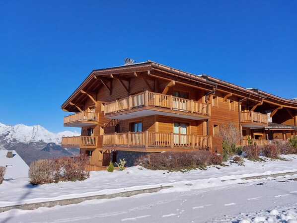 A wooden chalet with multiple balconies, set against a snowy mountain backdrop.