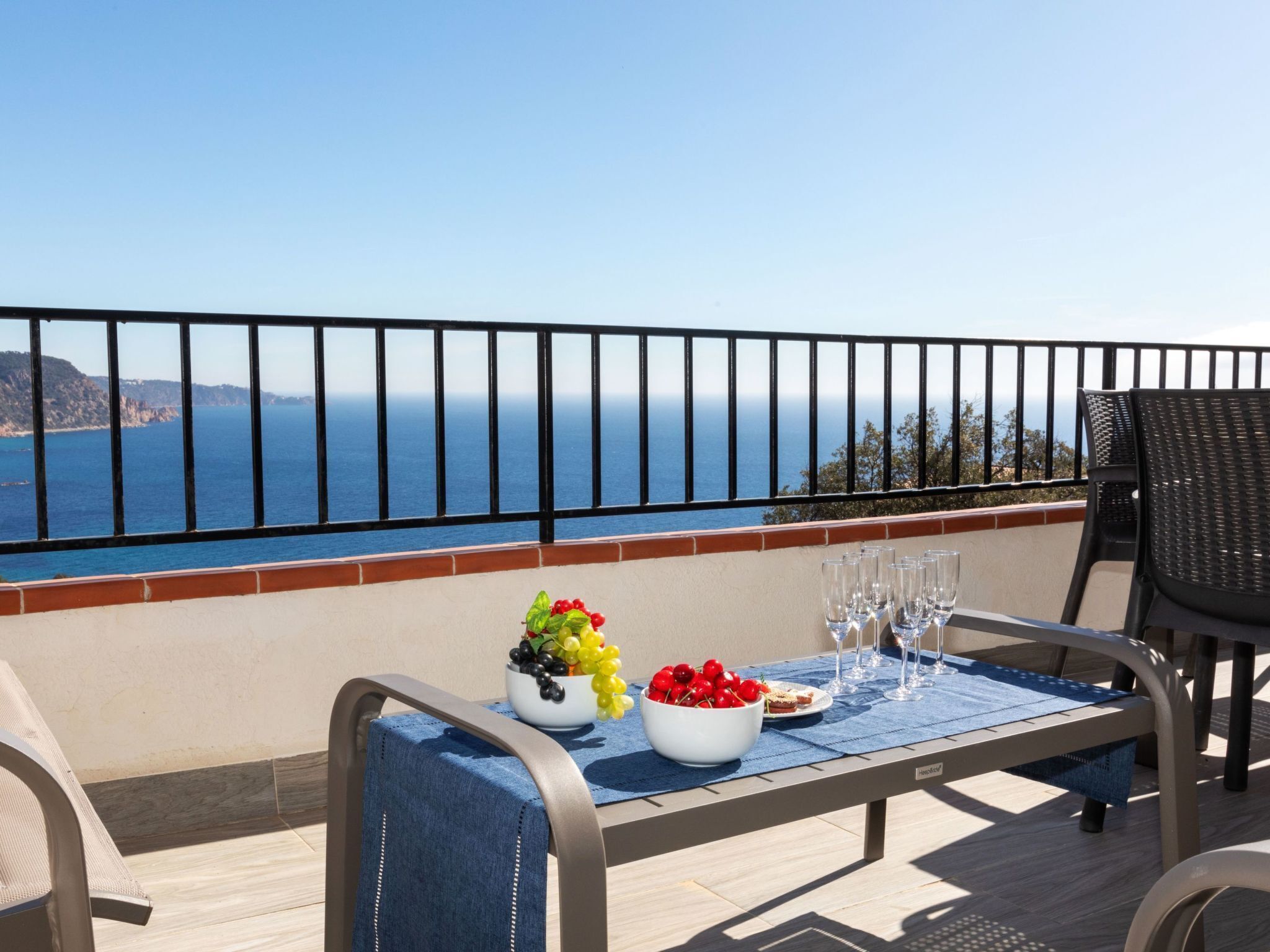 A balcony with a table set for dining, featuring a bowl of fruit and wine glasses, overlooking a coastal view.