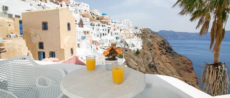 Una mesa con dos vasos de jugo de naranja y un florero con flores, con vista a un pueblo costero de edificios blancos y una palmera.