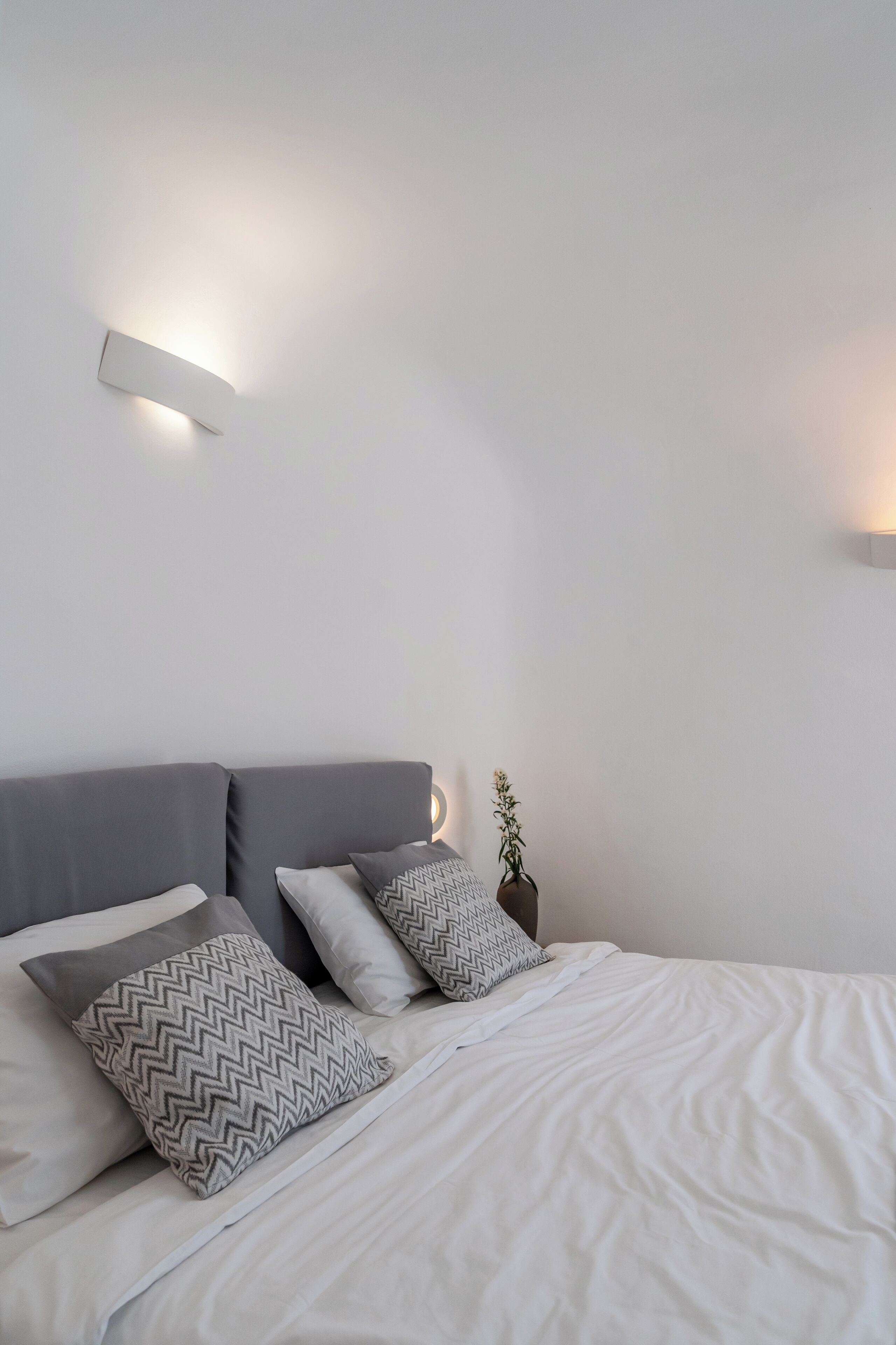 A neatly made bed with a gray headboard, white bedding, and patterned pillows. There are two wall-mounted lights on either side of the bed.