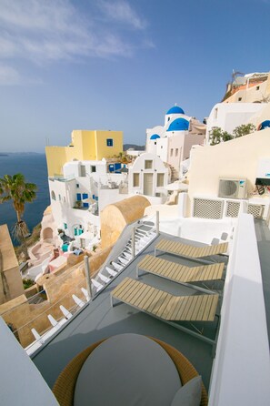 Una terraza en la azotea con vista al mar, edificios blancos con cúpulas azules y una palmera.