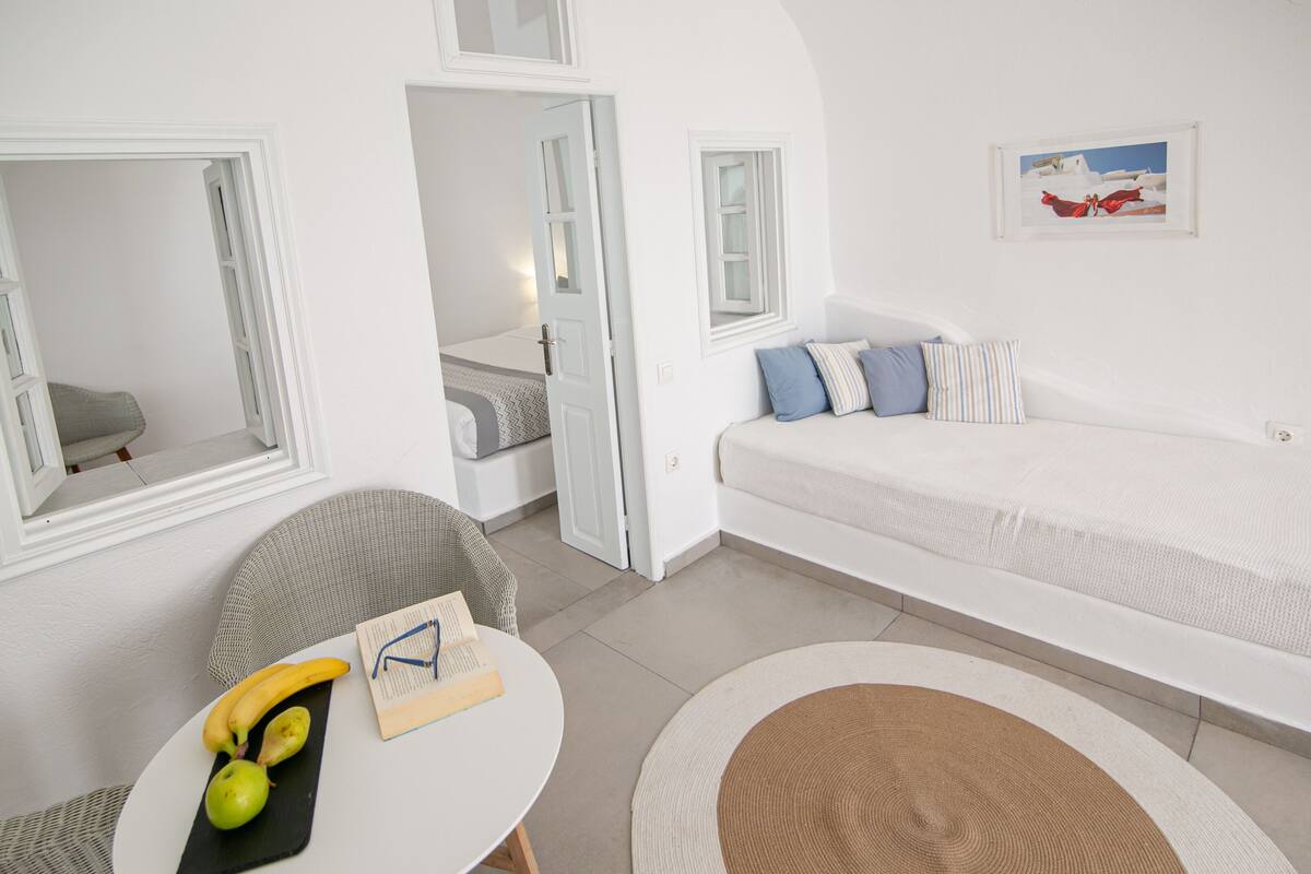 Ein modernes, minimalistisches Zimmer mit einem weißen Sofa, einem kleinen runden Tisch mit Früchten und einem Buch sowie Blick auf einen Balkon.