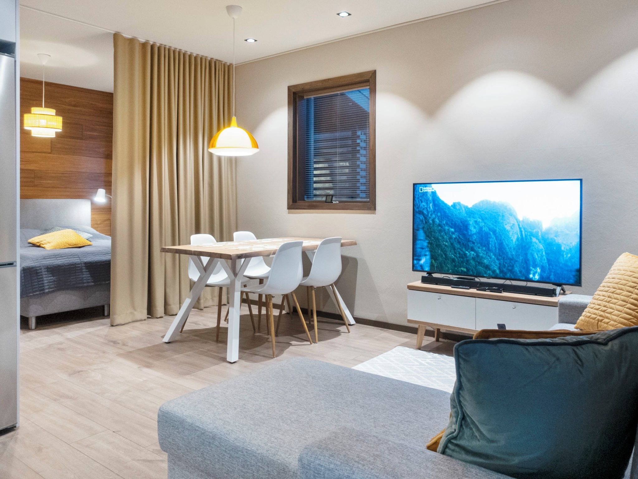 A modern living room with a sofa, a dining table with chairs, a television, and a window with blinds.