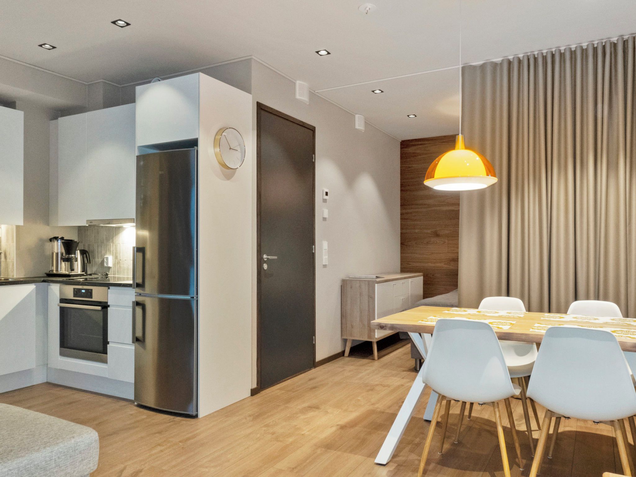 Une cuisine moderne dotée d’un four et d’un réfrigérateur encastrés, un coin repas avec une table et des chaises en bois, et une grande fenêtre avec des rideaux.