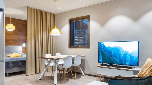 En moderne stue med et fladskærms-tv, et spisebord med stole, en sofa og et stort vindue med persienner.