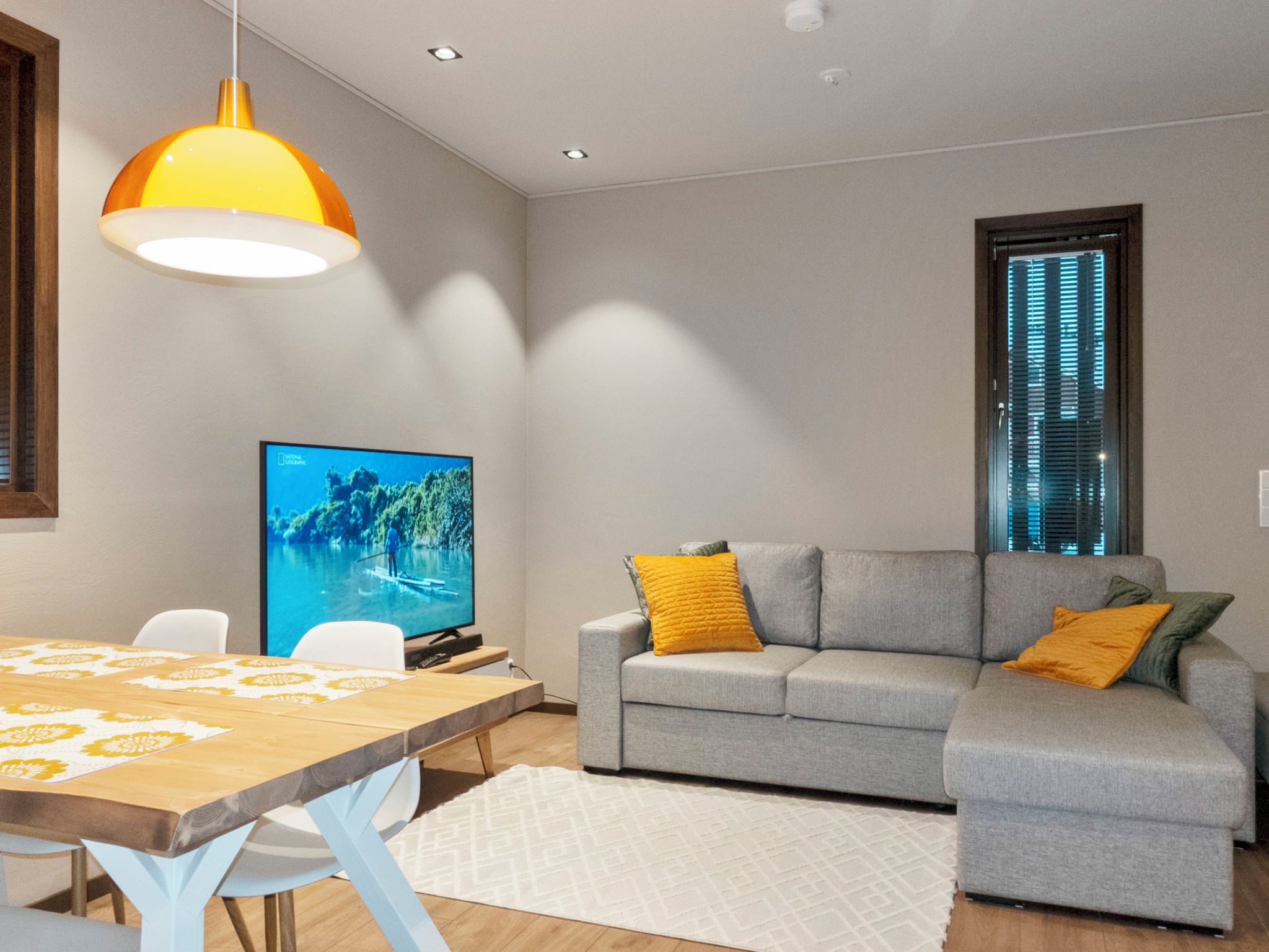 A modern living room with a sofa, a television, a dining table, and a window with blinds.
