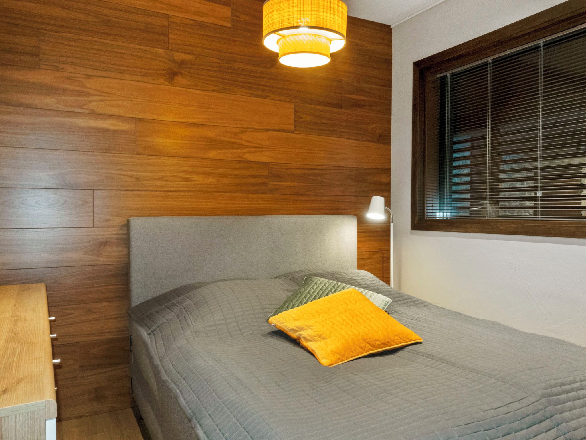 A bedroom with a wooden headboard, a bed with a grey quilt and a yellow pillow, a bedside lamp, and a window with blinds.