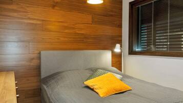 Une chambre à coucher avec une tête de lit en bois, un lit recouvert d’une couette grise et orné d’un oreiller jaune, une lampe de chevet et une fenêtre avec des stores.
