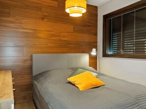 A bedroom with a wooden headboard, a bed with a grey quilt and a yellow pillow, a bedside lamp, and a window with blinds.