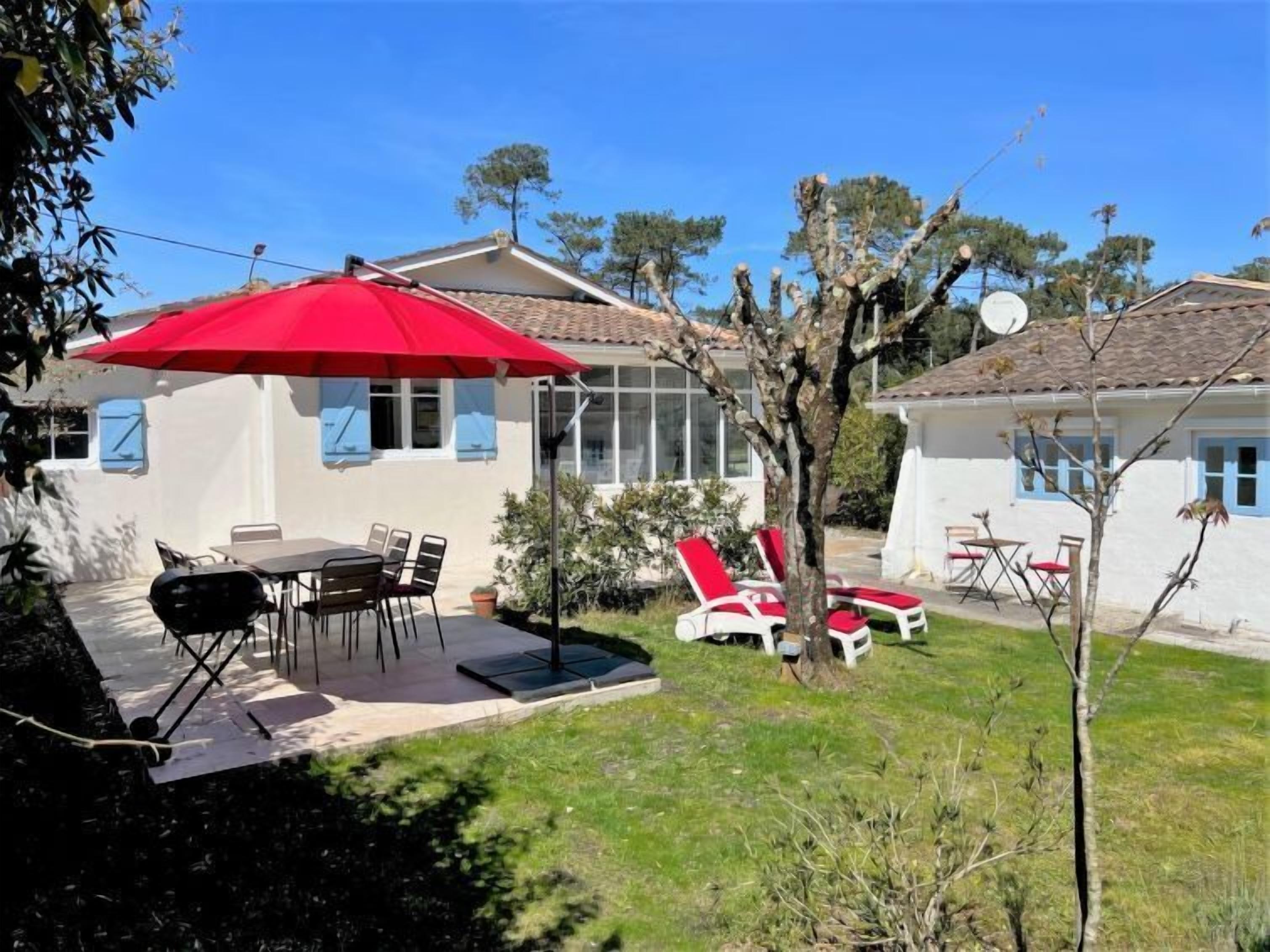 A patio with a table and chairs, a red umbrella, and a small garden with a tree and a white building in the background.