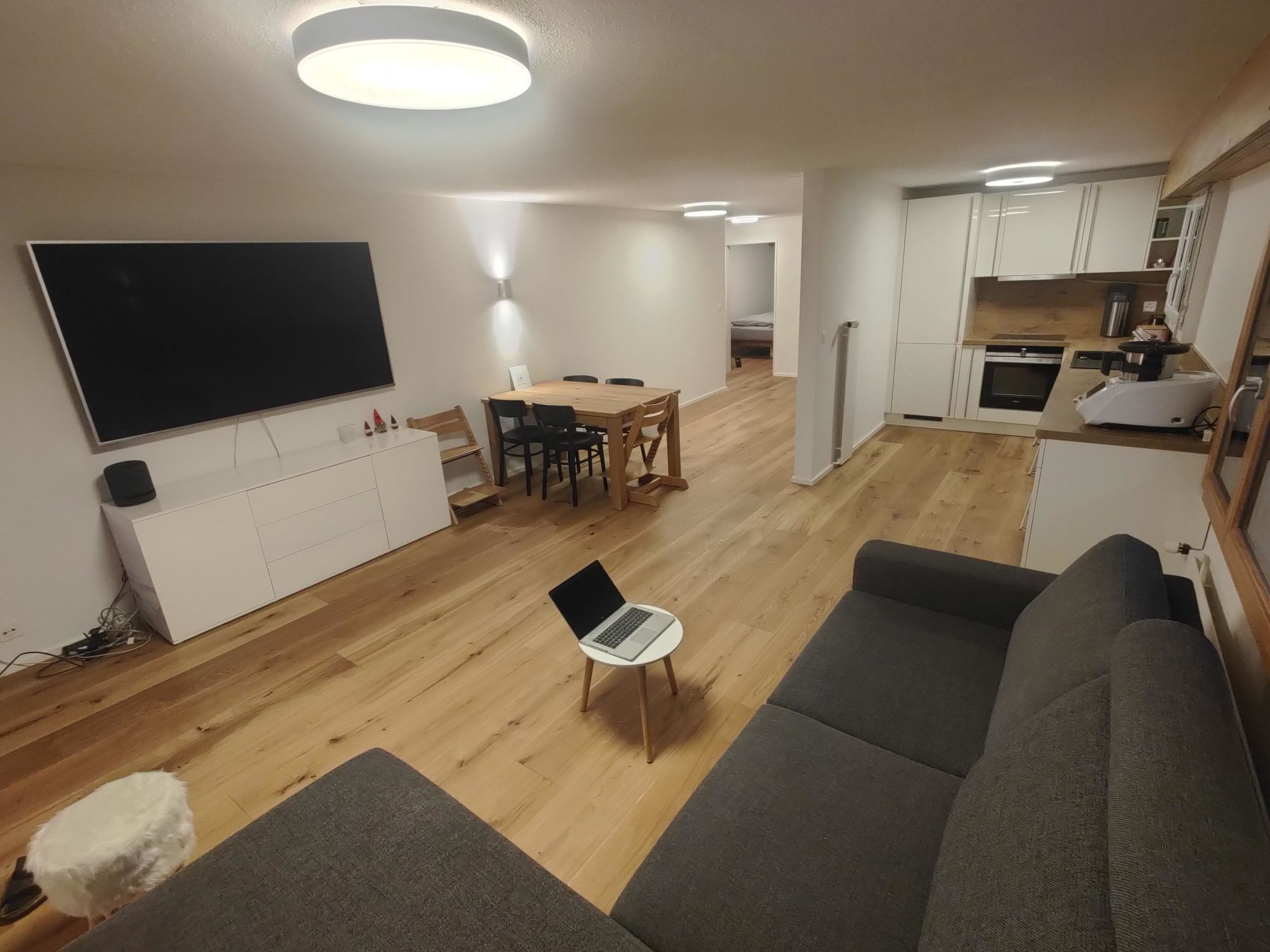 A modern living room with a flat-screen TV, a laptop on a small table, and a grey sofa.