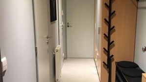 Un couloir étroit, avec une porte au fond, un radiateur au mur et un porte-manteau sur lequel est accroché un manteau.