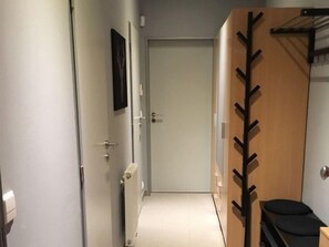 Un pasillo estrecho con una puerta al final, un radiador en la pared y un perchero con un abrigo colgado.