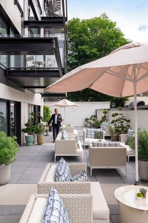A modern outdoor seating area with wicker furniture and large umbrellas, surrounded by potted plants and a building with large windows. - L-Avenue (Brussels)