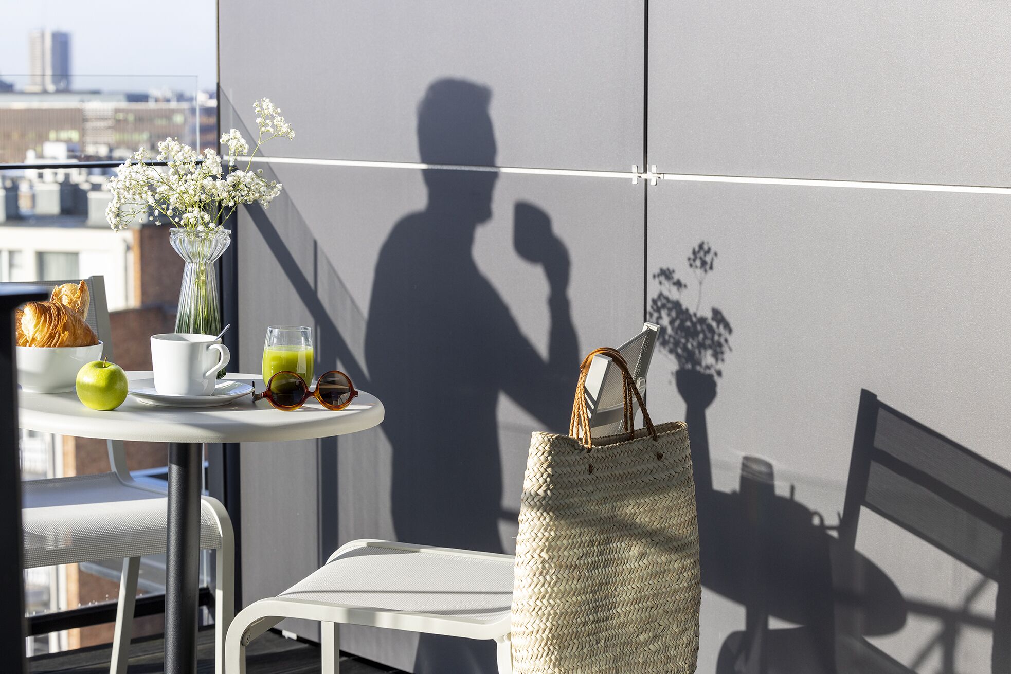 A balcony with a table set for breakfast, including a croissant, apple, coffee cup, and green juice. A woven bag hangs on the wall, and a shadow of a person is visible.