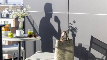 A balcony with a table set for breakfast, including a croissant, apple, coffee cup, and green juice. A woven bag hangs on the wall, and a shadow of a person is visible.