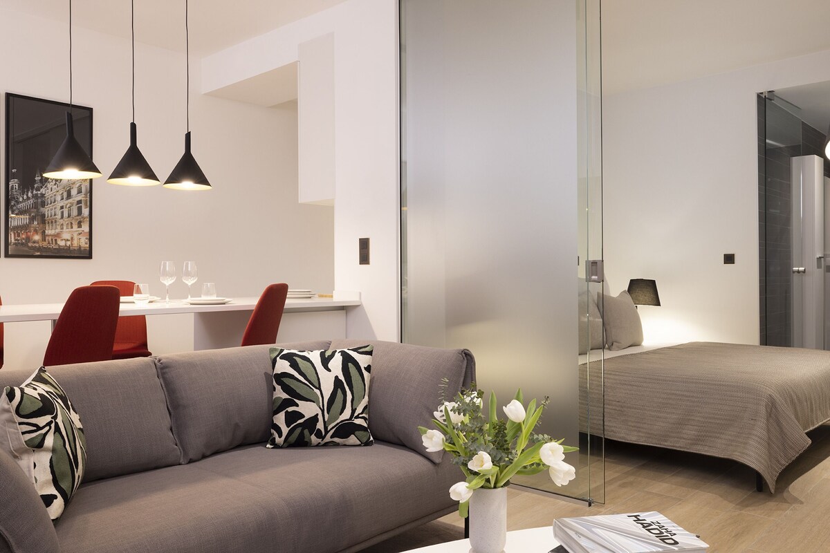 A modern hotel room with a sofa, a dining area with chairs and a table, a bed, and a glass partition.