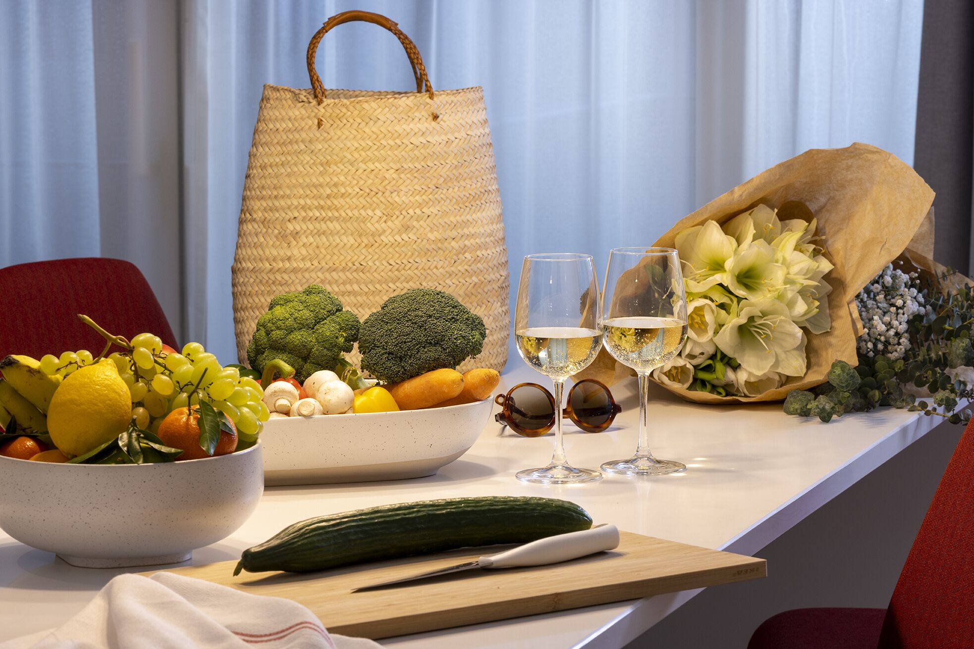 A kitchen counter with a bowl of fruit, a cucumber, a knife, a wine glass, and a bouquet of flowers.