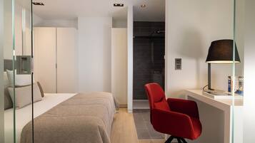 A modern hotel room with a bed, a red chair, a desk with a lamp, and a shower area.
