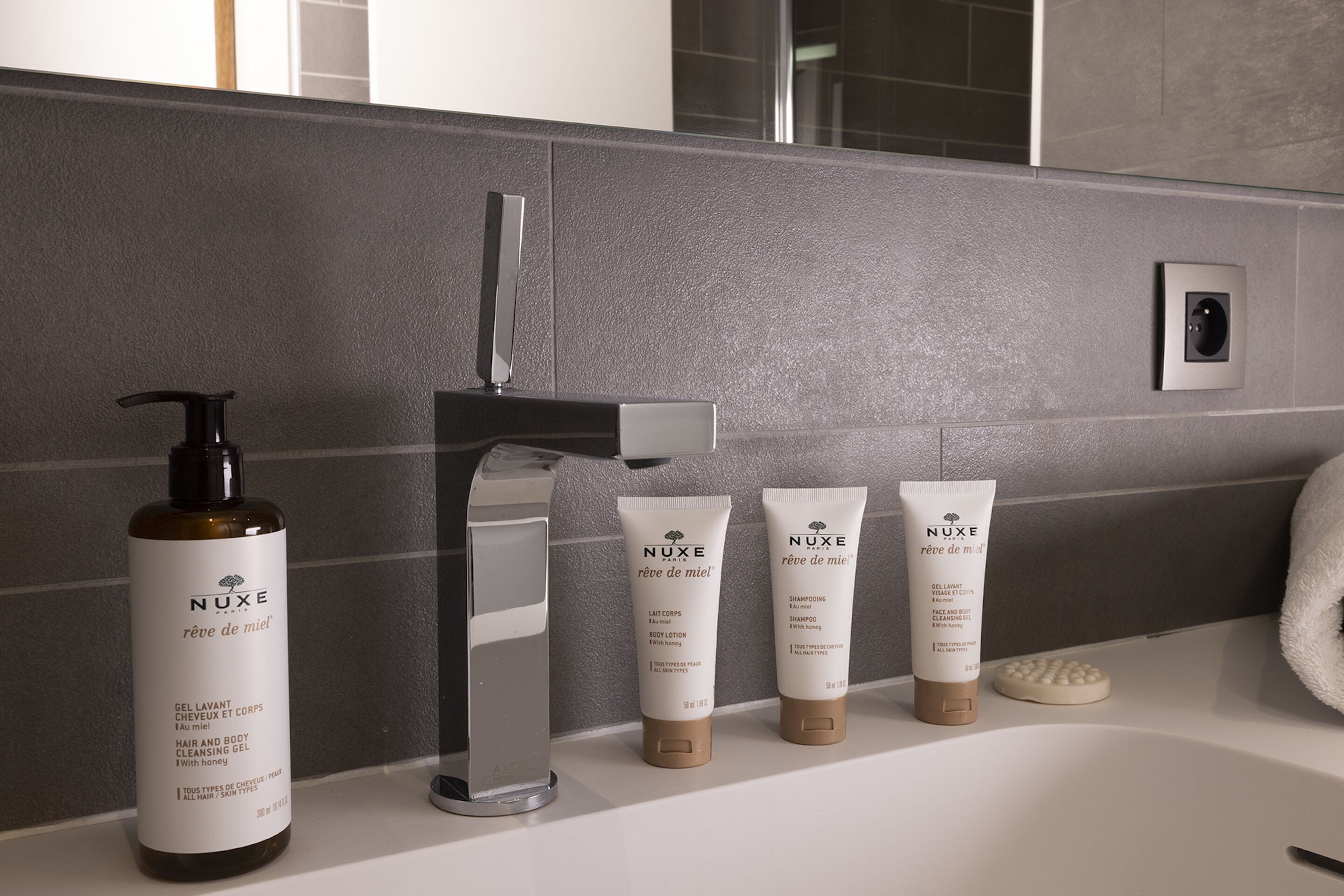 Une salle de bain avec un mur recouvert de carreaux gris, un robinet moderne et un flacon de gel nettoyant pour les cheveux et le corps de la marque Nuxe Paris.