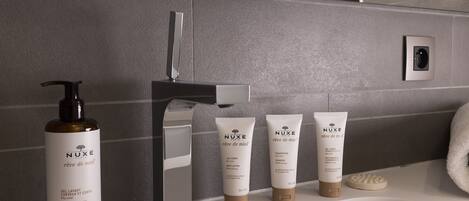 Une salle de bain avec un mur recouvert de carreaux gris, un robinet moderne et un flacon de gel nettoyant pour les cheveux et le corps de la marque Nuxe Paris.